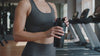 A woman in athletic wear holding a black shaker bottle while standing in front of treadmills in a gym.